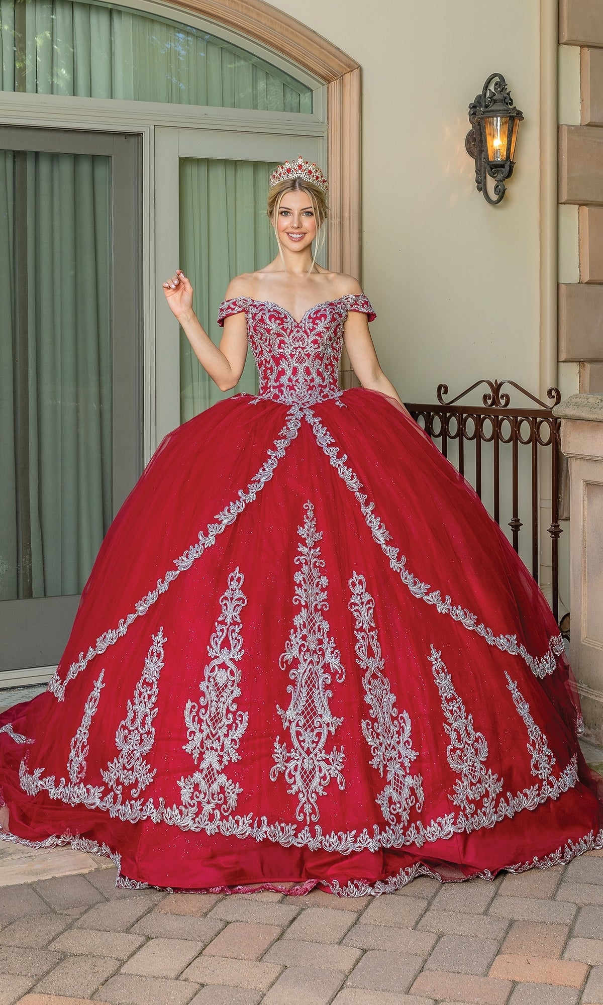 A young woman poses in front of large windows wearing the Red Off-the-Shoulder Quinceanera Dress 1662—a burgundy ball gown with a sweetheart neckline, silver embroidery, and a tiara—smiling outside by a lamp.
